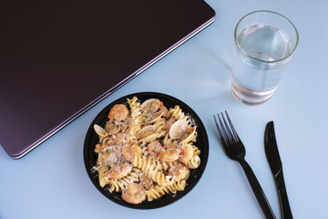 Seafood pasta business lunch at workplace. Open lunch box, closed laptop, knife and fork, glass of water on blue background.