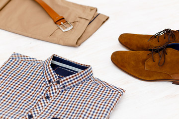 Beige pants, plaid shirt, brown suede shoes and leather belt. Overhead view of men's casual outfit on white wooden background.