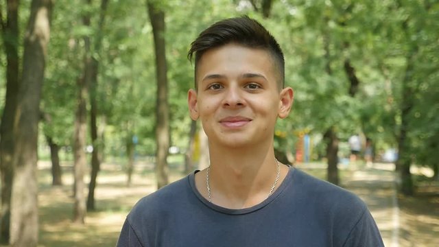  Portrait of a young brunet man with a short haircut  standing and smiling cheery in a green park ull of leafy trees on a sunny day 