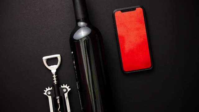 Red wine bottle, corkscrew and modern smartphone with red wallpeper on screen on black matte background. Top view with copy space