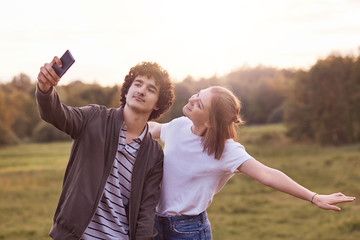Horizontal shot of positive best friends pose in smart phone for making selfie, enjoy spare time outside, being addicted to modern technologies, embrace each other. Teenagers, lifestyle concept