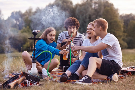 Teenagers have fun. Joyful girls and boys spend weekend outdoor on picnic, smoke hookah, clink bottles with energetic drinks, celebrate starting holidays. Friendship, youth and lifestyle concept