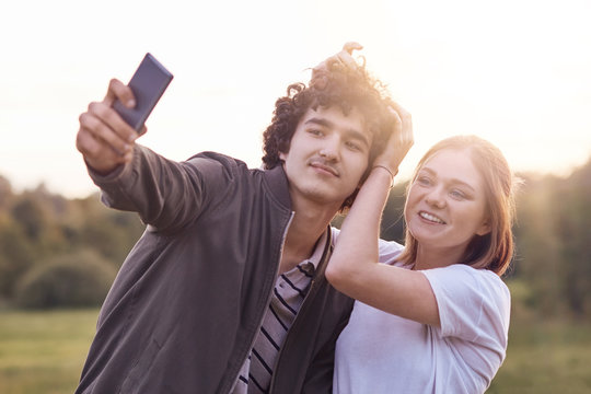 Funny curly male youngster and his girlfriend pose for making selfie portrait against blurred nature background, have positive expressions. Couple in love use modern cell phone for entertainment