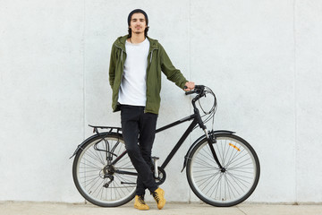 Isolated shot of stylish hipster male teenager dressed in stylish outfit and sneakers, stands near sport bicycle, enjoys recreation time alone, advertises his bike, poses over white concrete wall. - Powered by Adobe