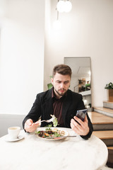 Man wearing a suit and a beard uses a smartphone in a cozy restaurant.Portrait of a business man sitting in a restaurant with salad and a cup of coffee on the table and using a smartphone.