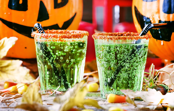 Halloween composition with festive green drink and smiling drinking pumpkins, with sweet corn, straw and fallen leaves on dark background, selective focus