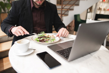 Business man eating breakfast in a restaurant. Plate of salad, a laptop, a cup of coffee, a smartphone on the table in the restaurant. Man eats a plate salad with a fork and a knife.
