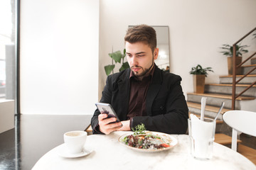 Portrait of a business man sitting in a restaurant with salad and a cup of coffee on the table and using a smartphone. Man wearing a suit and a beard uses a smartphone in a cozy restaurant.