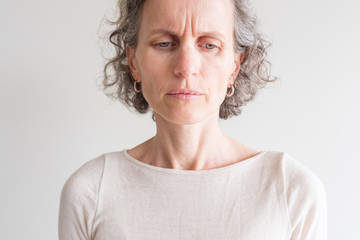 Close up head and shoulders view of middle aged woman with grey hair and cream top frowning and...