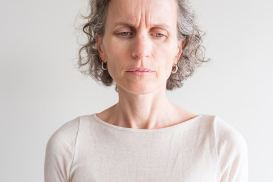 Close up head and shoulders view of middle aged woman with grey hair and cream top frowning and showing wrinkles (selective focus)