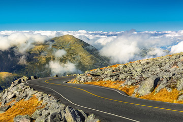 Winding road descending from Mount Washington, NH on a sunny autumn afternoon. Mount Jefferson peak stands above a thick layer of fluffy clouds.