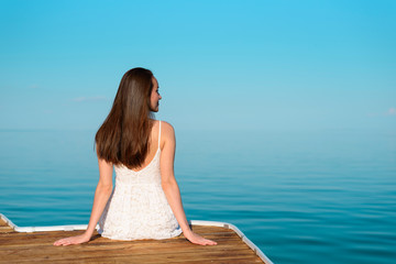 A young woman in a white dress sitting on a wooden pier looking away, against the blue sea