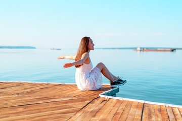 Fototapeta na wymiar Young girl in white dress sitting on a wooden pier with open hands looking into the distance of the sea, freedom, clean air, dreams