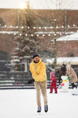 Portrait of joyful Scandinavian bearded man in yellow jacket, beige trousers on ice rink, outdoors in snowy winter day, Christmas tree on background. Weekend activities outdoor in cold weather