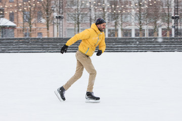 Portrait of bearded man in yellow jacket, beige trousers, black hat on ice rink, outdoors in snowy winter day, side view. Handsome man shooting in motion on ice rink © DimaBerlin