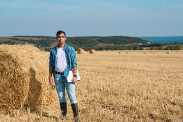 Naklejka na ściany i meble Young farmer in jeans, white t-shirt, blue shirt, rubber boots in the field with haystacks with tablet and notebook, copy space, agricultural industry, rural business concept