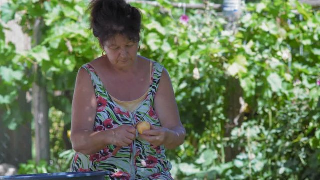 Elderly Woman Takes an Apple from an Iron Bowl and Cleans it with a Peel Knife