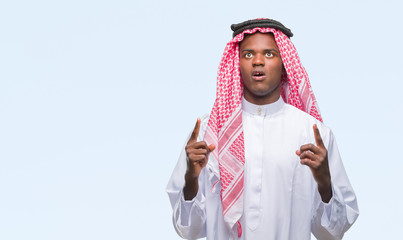 Young arabic african man wearing traditional keffiyeh over isolated background amazed and surprised looking up and pointing with fingers and raised arms.