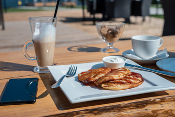 Deadpan image of pancake with honey, sour cream on wooden table with fork, spoon, smartphone and Cup of tea