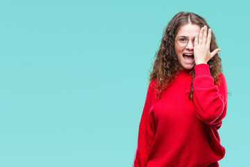 Beautiful brunette curly hair young girl wearing glasses and winter sweater over isolated background covering one eye with hand with confident smile on face and surprise emotion.