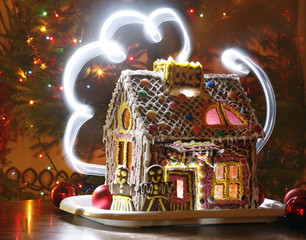 Beautiful well decorated gingerbread house in front of are gingerbread man and woman on white tray in home living room in front of the decorated christmas tree. Candy windows glowing, dark night.