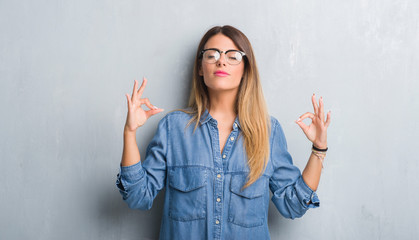 Young adult woman over grunge grey wall wearing glasses relax and smiling with eyes closed doing meditation gesture with fingers. Yoga concept.