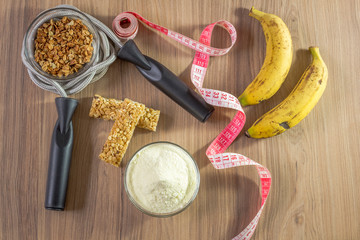 Fitness concept with dumbbells skipping rope measure tape whey protein and bananas on wooden table background.