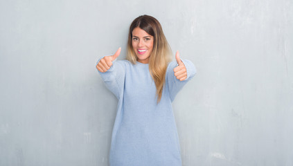 Young adult woman over grey grunge wall wearing winter outfit approving doing positive gesture with hand, thumbs up smiling and happy for success. Looking at the camera, winner gesture.