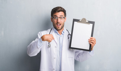 Handsome young doctor man over grey grunge wall holding clipboard with surprise face pointing finger to himself