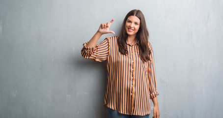 Young brunette woman over grunge grey wall smiling and confident gesturing with hand doing size sign with fingers while looking and the camera. Measure concept.