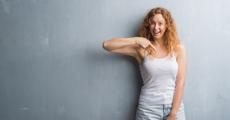 Young redhead woman over grey grunge wall with surprise face pointing finger to himself