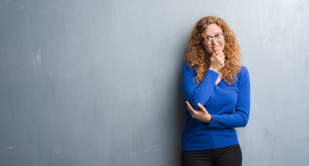 Fototapeta premium Young redhead woman over grey grunge wall looking confident at the camera with smile with crossed arms and hand raised on chin. Thinking positive.