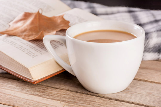 Coffee cup on retro wooden desk and open book with fallen dry leaf and blanket blurred in background. Autumn concept image. Close up, selective focus