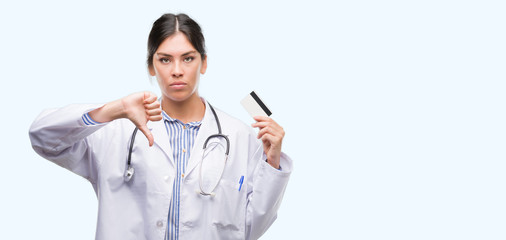 Young hispanic doctor woman holding credit card with angry face, negative sign showing dislike with thumbs down, rejection concept