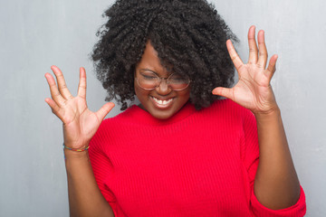 Young african american woman over grey grunge wall wearing winter sweater and glasses celebrating mad and crazy for success with arms raised and closed eyes screaming excited. Winner concept