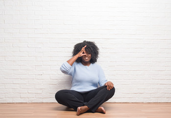 Young african american woman sitting on the floor at home doing ok gesture with hand smiling, eye looking through fingers with happy face.