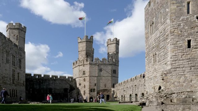 Visitors tour Caernarfon Castle court yard grounds as flags wave from twin towers against white cloud in a blue sky