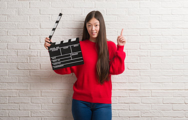 Young Chinese woman over white brick wall holding clapboard surprised with an idea or question...