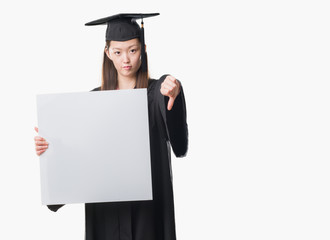 Young Chinese woman wearing graduate uniform holding banner with angry face, negative sign showing dislike with thumbs down, rejection concept