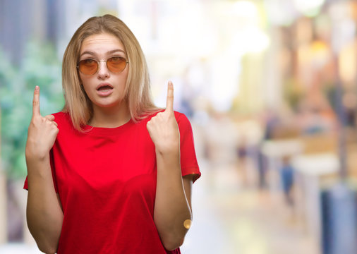 Young caucasian woman wearing sunglasses over isolated background amazed and surprised looking up and pointing with fingers and raised arms.