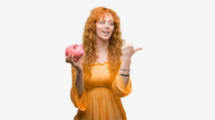 Young redhead woman holding piggy bank pointing and showing with thumb up to the side with happy face smiling