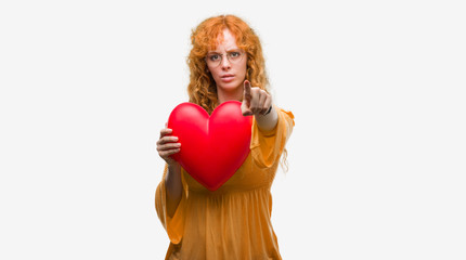 Young redhead woman in love holding red heart pointing with finger to the camera and to you, hand sign, positive and confident gesture from the front