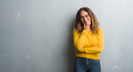 Middle age hispanic woman over grey wall wearing glasses looking confident at the camera with smile with crossed arms and hand raised on chin. Thinking positive.