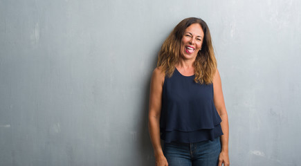 Fototapeta premium Middle age hispanic woman standing over grey grunge wall sticking tongue out happy with funny expression. Emotion concept.