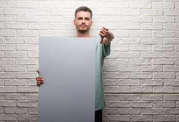Young adult man over brick wall holding banner with angry face, negative sign showing dislike with...