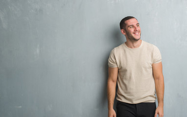 Young caucasian man over grey grunge wall looking away to side with smile on face, natural expression. Laughing confident.