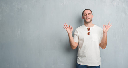 Young caucasian man over grey grunge wall wearing sunglasses relax and smiling with eyes closed doing meditation gesture with fingers. Yoga concept.