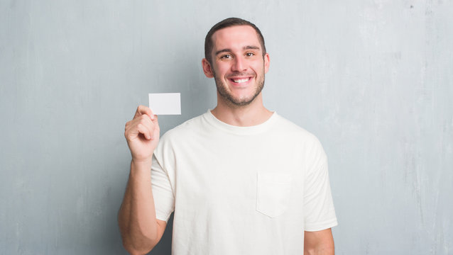 Young caucasian man over grey grunge wall showing blank visit card with a happy face standing and smiling with a confident smile showing teeth