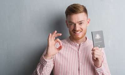 Young redhead man over grey grunge wall holding passport of Australia doing ok sign with fingers,...
