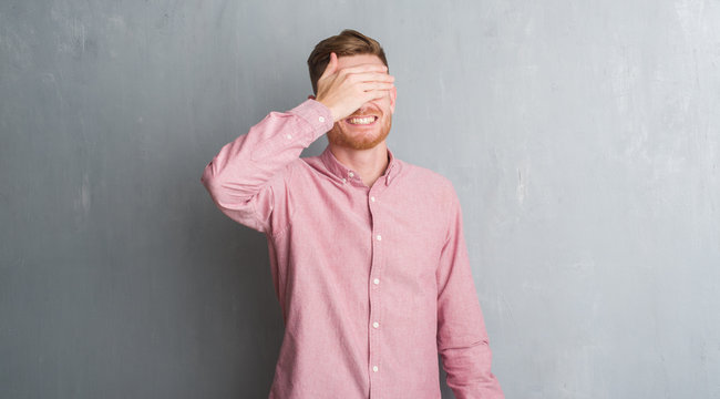 Young redhead man over grey grunge wall wearing pink shirt smiling and laughing with hand on face covering eyes for surprise. Blind concept.